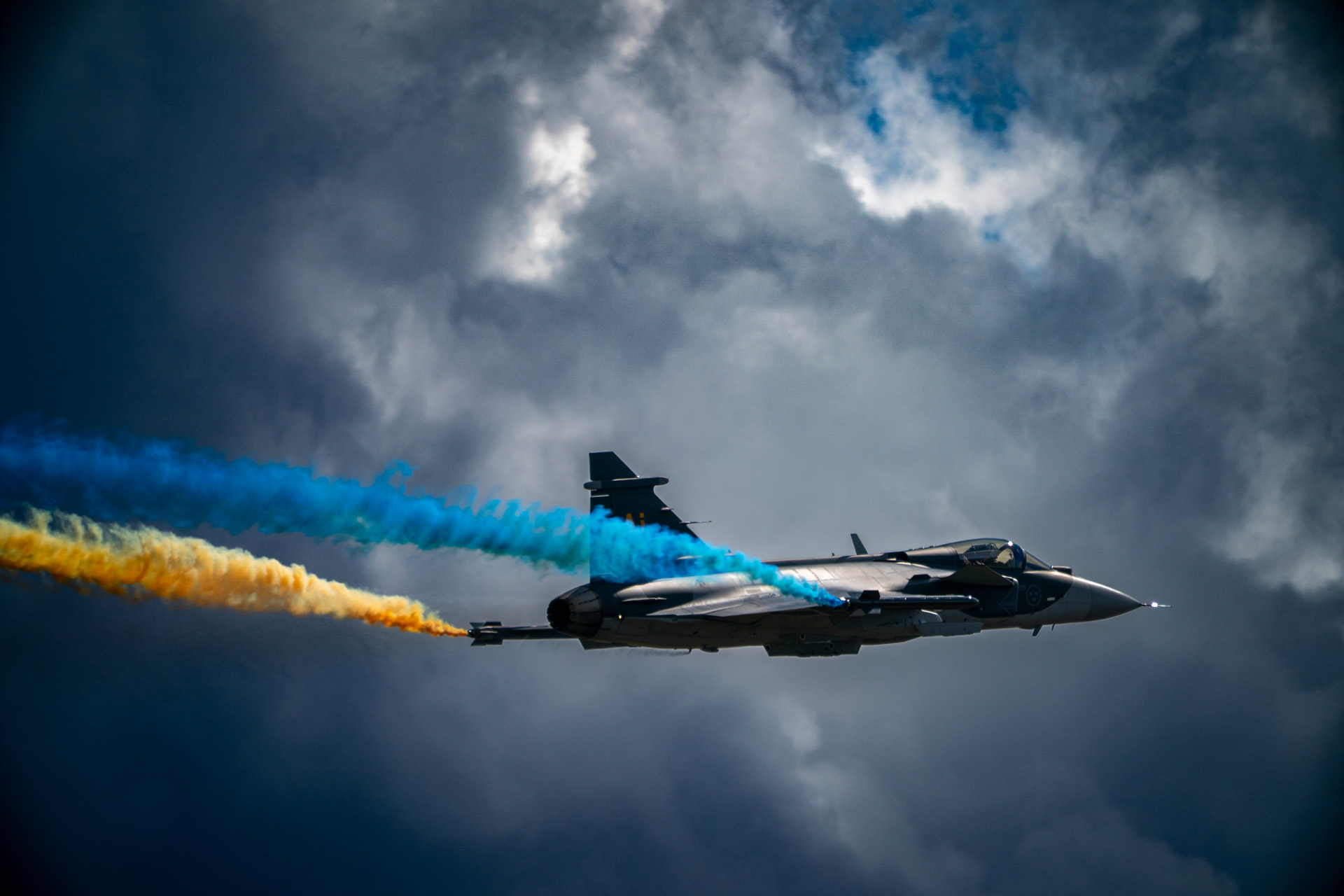 Swedish Air Force Gripen flying with blue and yellow Sanders Smoke against dramatic clouds