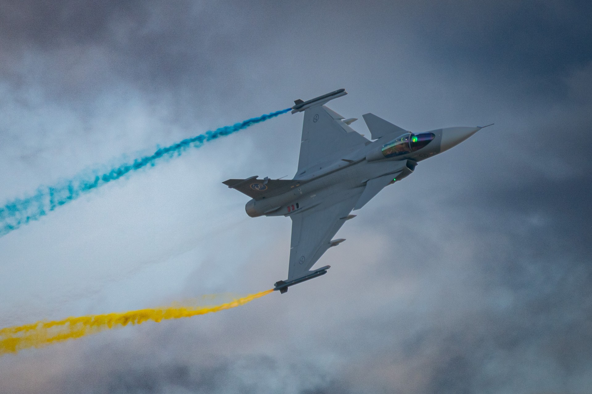 Blue and yellow aerial smoke crossing the sky behind a Swedish Gripen aircraft